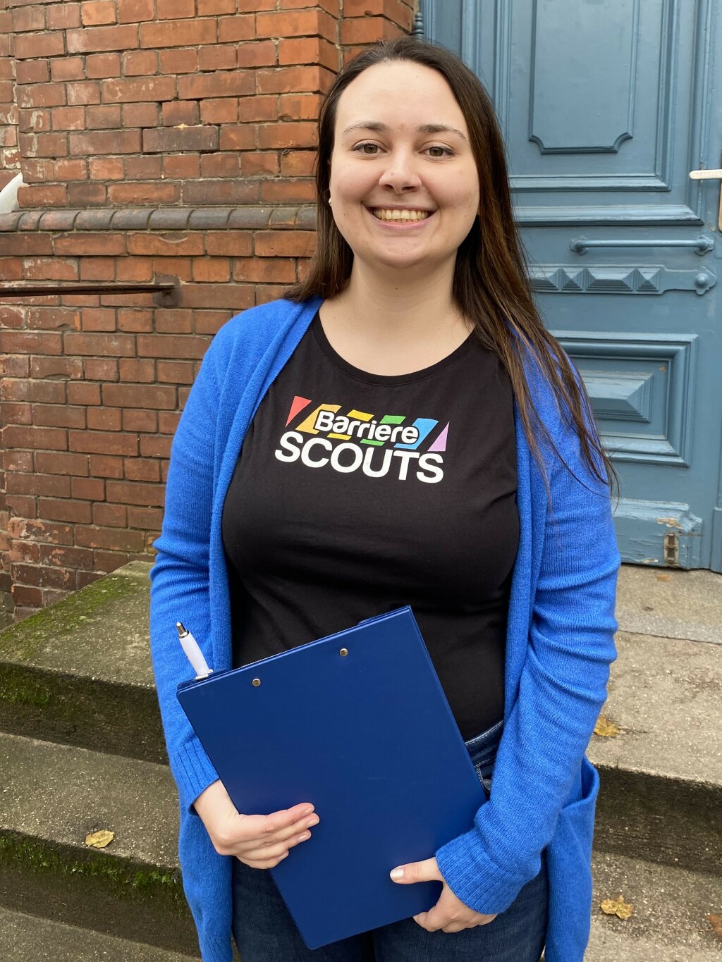 Sharon Maple lächelt in die Kamera. Sie trägt ein T-Shirt mit dem Aufdruck Barriere Scouts und hält ein blaues Klemmbrett mit Stift in der Hand. Sie steht vor einem Eingang mit Treppen.